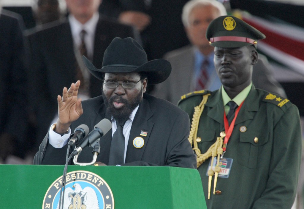 H.E. President Salva Kiir Mayardit speaking at the green presidential podium with the presidential seal, microphones, and a military officer in uniform at an outdoor state event